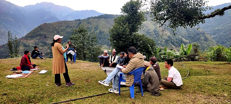 A facilitator leads an outdoor community session on a hillside in Dailekh, with participants seated on mats and chairs and green mountains behind